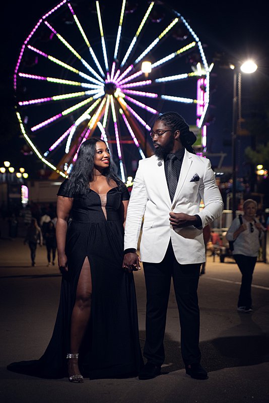 Black man with locs and a white blazer and black woman with long hair, a long black dress and red lipstick in at nc state fair engagement session