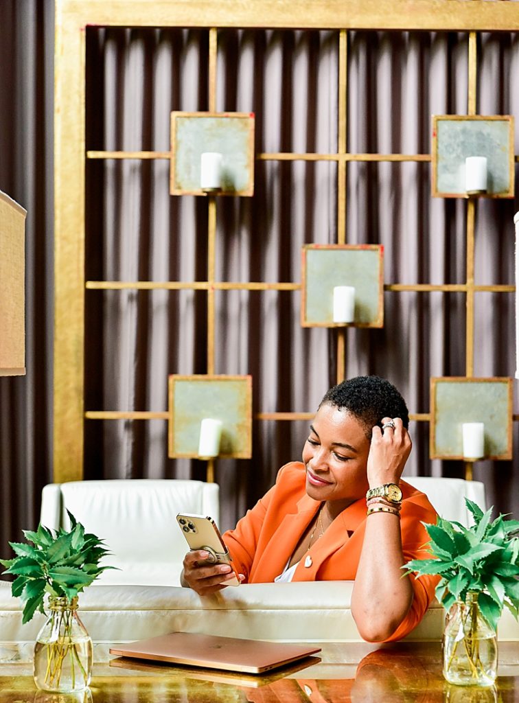 Fashionable Black business woman wearing orange blazer, while working on laptop and cell phone in the lobby of Proximity Hotel, Greensboro NC