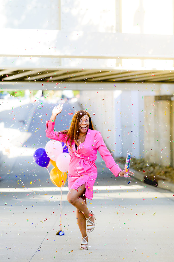 Black woman in pink dress standing outside in front of balloons with confetti coming down. Five Must Have Photos For Your Brand.